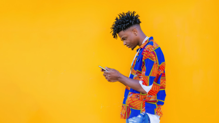 A man african print shirt stands his body the right looking at his phone against yellow background