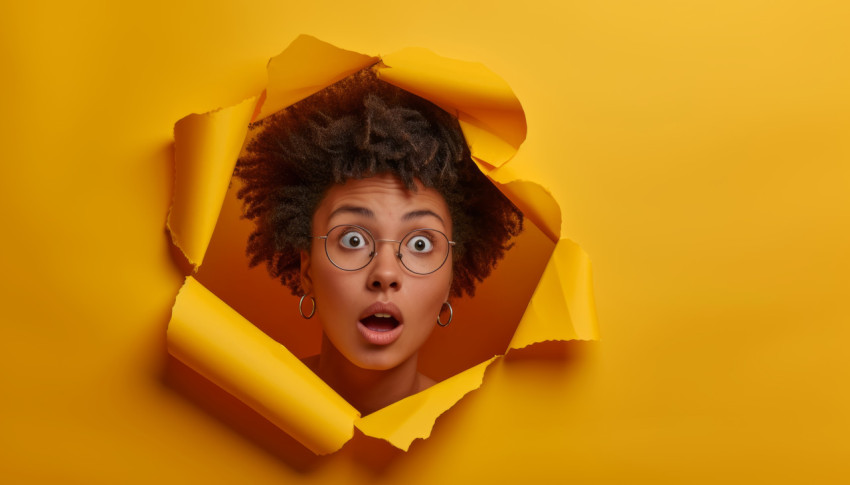 Surprised young woman emerges through yellow paper hole expressing amazement and curiosity in a whimsical setting