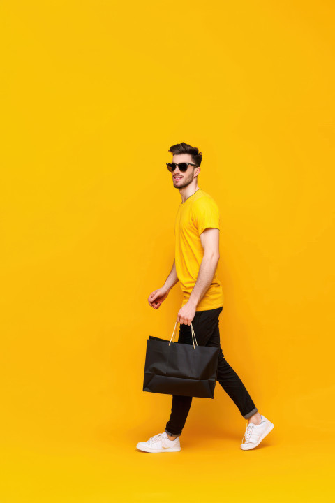 A man in sunglasses and a yellow t shirt carrying a black shopping bag representing black friday sales